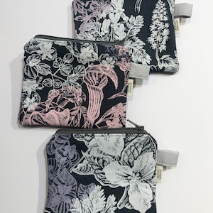 Könnte beinhalten: Drei kleine, quadratische Münzbörsen mit floralen Mustern. Die Geldbörsen haben einen schwarzen Hintergrund mit rosa, weißen und lila botanischen Designs. Jede Geldbörse hat einen grauen Reißverschluss und ein kleines graues Stoffetikett.