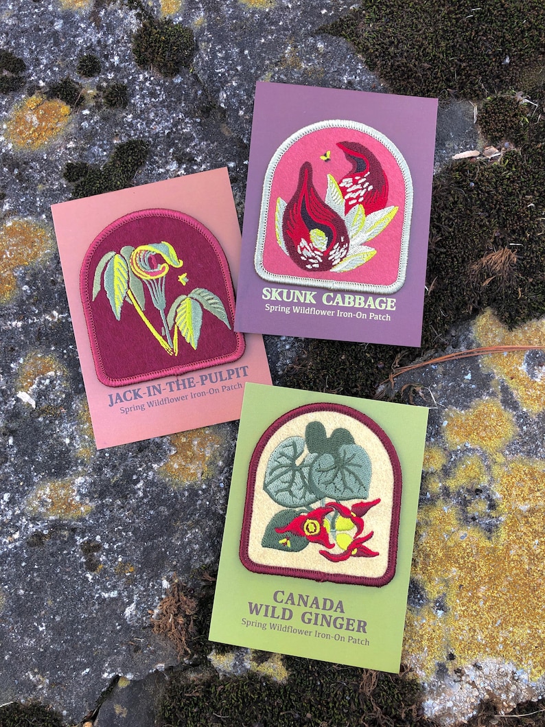 May include: Three iron-on patches featuring illustrations of spring wildflowers. The patches are on cards with the names "Jack-in-the-Pulpit", "Skunk Cabbage", and "Canada Wild Ginger".