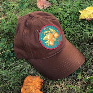 May include: A brown baseball cap with a circular embroidered patch featuring a green background, orange fruit, and yellow leaves.