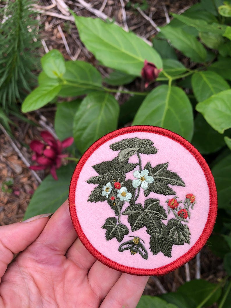 May include: A circular embroidered patch featuring a light pink background with a red stitched border. The design depicts a botanical illustration of green leaves, white flowers, small red berries, and a small bee in the lower section.