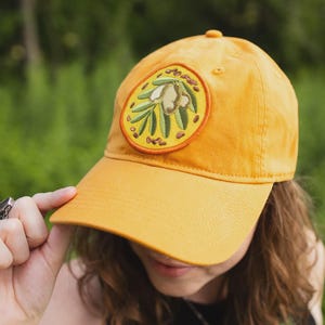 May include: A mustard yellow baseball cap featuring a circular embroidered patch on the front. The patch displays a detailed illustration of green olive branches with olives and small brown accents, all bordered by a vibrant orange stitched edge.