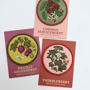 May include: Three circular embroidered iron-on patches featuring botanical illustrations of wild fruits. The patches depict Common Serviceberry, Prickly Gooseberry, and Thimbleberry, each mounted on a rectangular card with corresponding text labels.