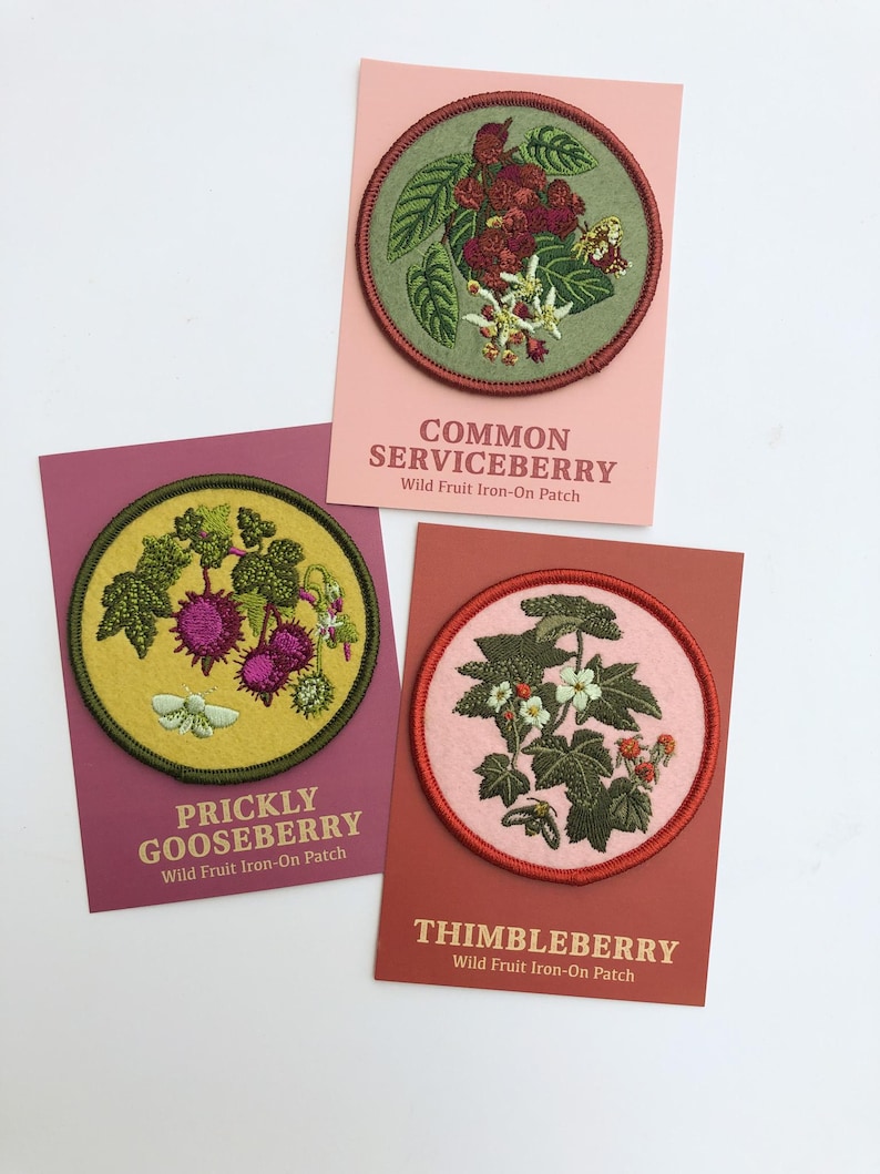 May include: Three circular embroidered iron-on patches featuring botanical illustrations of wild fruits. The patches depict Common Serviceberry, Prickly Gooseberry, and Thimbleberry, each mounted on a rectangular card with corresponding text labels.