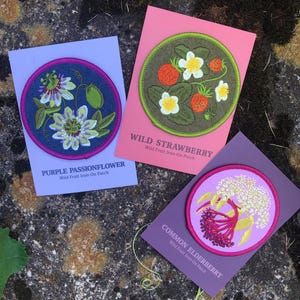 May include: Three embroidered patches featuring floral designs. One patch displays a purple passionflower, another shows wild strawberries, and the third depicts common elderberry. Each patch is on a colored card with the plant name and "Wild Fruit Iron-On Patch" text.