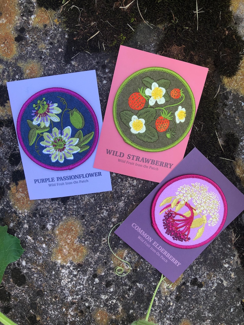 May include: Three embroidered patches featuring floral designs. One patch displays a purple passionflower, another shows wild strawberries, and the third depicts common elderberry. Each patch is on a colored card with the plant name and "Wild Fruit Iron-On Patch" text.