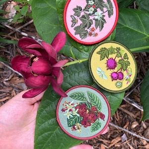 May include: Three circular embroidered patches featuring botanical designs. The top patch is light pink with green leaves and white flowers. The middle patch is yellow with green leaves and purple berries. The bottom patch is sage green with red berries.