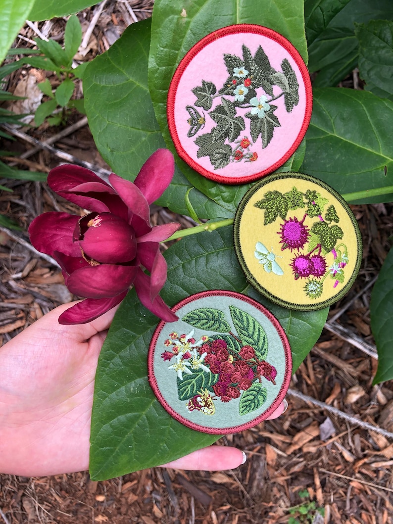 May include: Three circular embroidered patches featuring botanical designs. The top patch is light pink with green leaves and white flowers. The middle patch is yellow with green leaves and purple berries. The bottom patch is sage green with red berries.