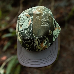 Puede incluir: Gorra de cinco paneles con un estampado floral en tonos verdes, naranjas y blancos. La visera es de color gris liso. La gorra está hecha de una tela duradera, adecuada para actividades al aire libre.