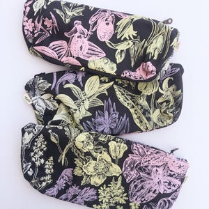Könnte beinhalten: Drei schwarze Reißverschlusstaschen mit einem Blumenmuster in Rosa, Gelb und Grün. Die Taschen haben eine gebogene Form und sind vertikal angeordnet. Das florale Design umfasst verschiedene Blumenarten und Blätter vor schwarzem Hintergrund.