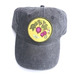 May include: A charcoal grey baseball cap featuring a circular patch on the front. The patch has a yellow background with an embroidered design of green leaves, pink spiky fruits, and a small light green moth, all bordered by a dark green edge.