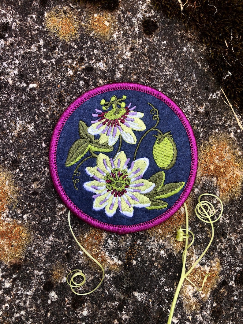 May include: A circular embroidered patch featuring a passion flower design. The patch has a navy blue background with a magenta border. The flower is depicted in shades of yellow, green, and purple, with green leaves and a green fruit. The patch is likely for clothing or accessories.