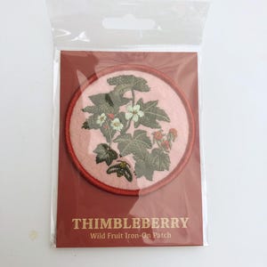 May include: A circular iron-on patch featuring an embroidered thimbleberry plant with green leaves, white flowers, and small red berries on a light pink background, enclosed in a red border, packaged with a card labeled THIMBLEBERRY Wild Fruit Iron-On Patch.