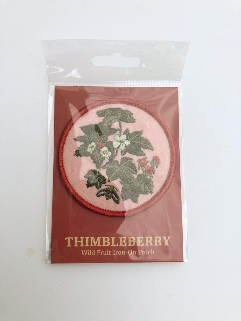 May include: A circular iron-on patch featuring an embroidered thimbleberry plant with green leaves, white flowers, and small red berries on a light pink background, enclosed in a red border, packaged with a card labeled THIMBLEBERRY Wild Fruit Iron-On Patch.
