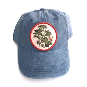 May include: A blue denim baseball cap featuring a circular patch on the front. The patch has a light pink background with a red border and displays an embroidered botanical illustration of green leaves, white flowers, and small red berries.
