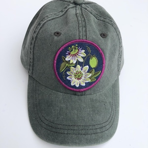 May include: A dark green baseball cap with a circular embroidered patch. The patch features a detailed floral design with white and purple flowers, green leaves, and a green fruit, set against a dark blue background with a pink border. The cap has a curved brim.