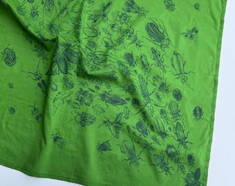 Puede incluir: Un cuadrado de tela verde brillante con un estampado repetitivo en verde oscuro de varias ilustraciones de escarabajos e insectos dispersas por la superficie. El tejido tiene una textura suave y plegada con bordes dobladillados visibles.