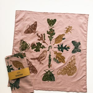 May include: A dusty rose bandana with a circular design of insects, leaves, and a bird in shades of brown and green. A matching folded bandana with a label that reads "Bandana" is also present.