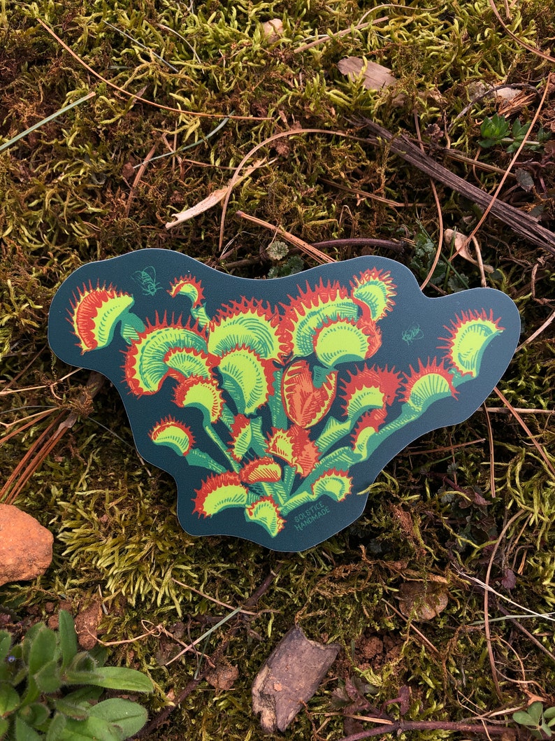 May include: A sticker featuring a detailed illustration of a Venus flytrap plant with red, green, and yellow colors on a dark blue background. The sticker has the text "SORTRICE HANDMADE" at the bottom.