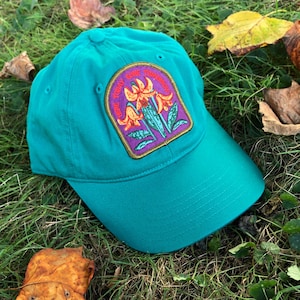 May include: A teal blue baseball cap with an embroidered patch featuring three orange lilies and the text "Here today gone tomorrow".