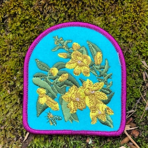May include: Embroidered patch featuring a floral design with yellow flowers and green leaves on a turquoise background. The patch has a purple border and a small bee. The patch is set against a mossy background.