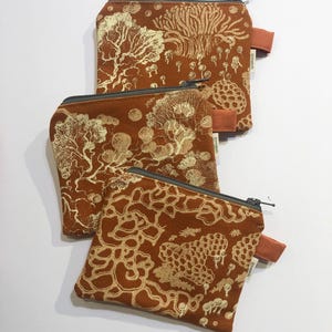 May include: Three small, zippered pouches in varying sizes, featuring a repeating gold pattern on a rust-colored background. The pattern includes stylized trees and organic shapes. Each pouch has a brown fabric pull tab and a dark gray zipper.
