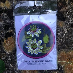 May include: A round, embroidered patch featuring a purple passionflower design with green leaves and a green fruit. The patch has a dark blue background with a pink border. Text at the bottom reads "PURPLE PASSIONFLOWER Wild Fruit Iron-On Patch."