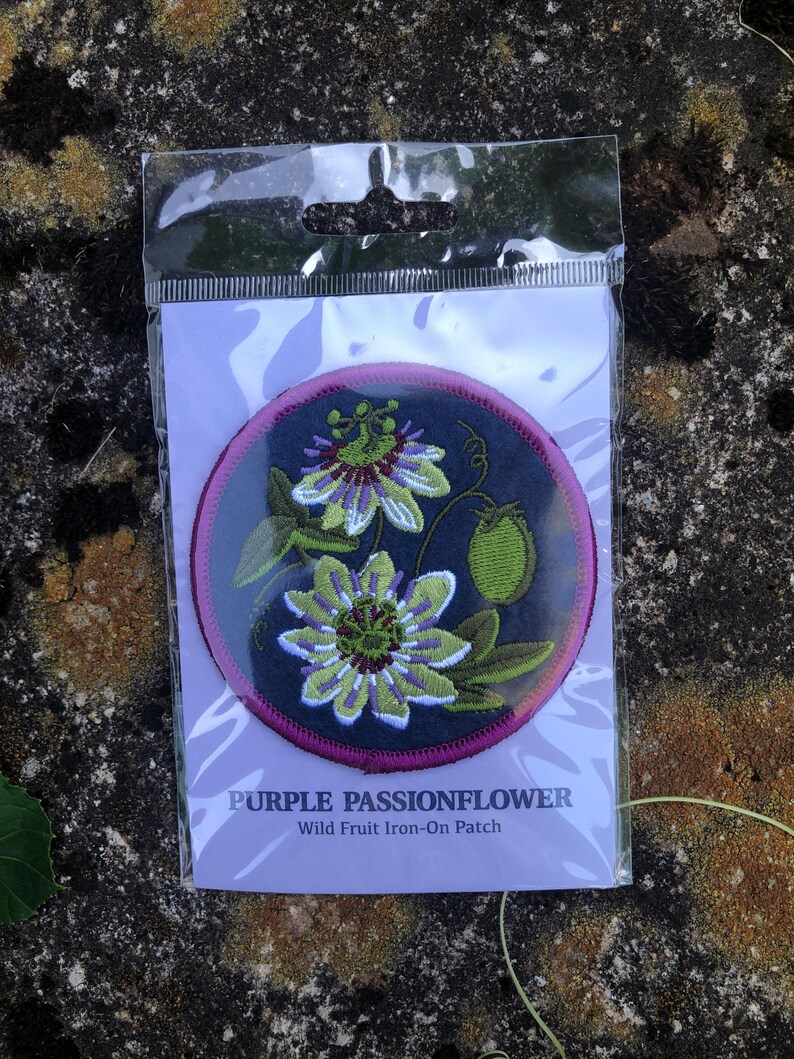 May include: A round, embroidered patch featuring a purple passionflower design with green leaves and a green fruit. The patch has a dark blue background with a pink border. Text at the bottom reads "PURPLE PASSIONFLOWER Wild Fruit Iron-On Patch."