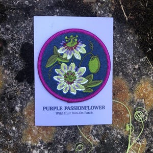 May include: A circular iron-on patch featuring a purple passionflower design. The patch has a dark blue background with a magenta border and green and white floral details. The text "PURPLE PASSIONFLOWER" and "Wild Fruit Iron-On Patch" are printed below the patch.