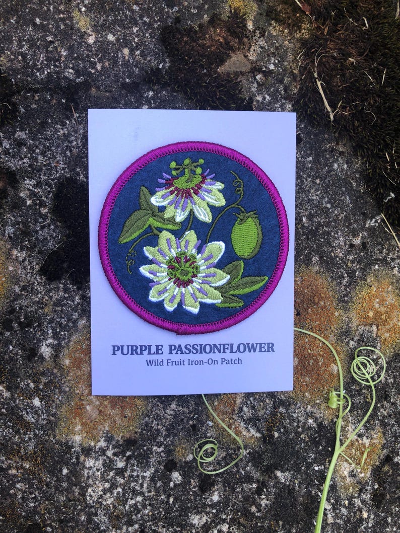 May include: A circular iron-on patch featuring a purple passionflower design. The patch has a dark blue background with a magenta border and green and white floral details. The text "PURPLE PASSIONFLOWER" and "Wild Fruit Iron-On Patch" are printed below the patch.