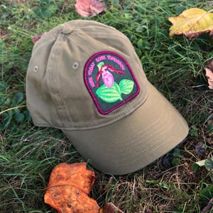 May include: A green baseball cap with a pink and green embroidered patch that says "Here Today Gone Tomorrow!" The patch features a pink flower with green leaves.