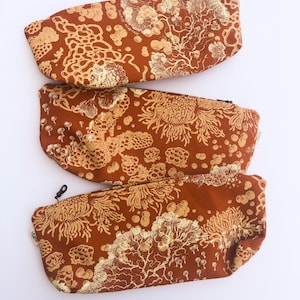 May include: Three rust-colored zippered pouches with a gold floral pattern. The pouches are rectangular with rounded ends. The gold pattern includes stylized flowers and organic shapes. Each pouch has a black zipper pull.