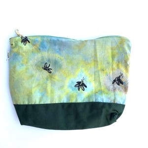 Könnte beinhalten: Eine Reißverschlusstasche mit grünem und blauem Batikmuster und dunkelgrünem Boden. Die Tasche hat einen goldenen Reißverschluss und schwarze Bienenillustrationen. Wahrscheinlich für Kosmetika oder kleine Gegenstände.