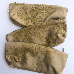 Könnte beinhalten: Drei beige Stoffbeutel mit einem sich wiederholenden olivgrünen Blatt- und Fruchtmuster. Jeder Beutel hat einen schwarzen Reißverschluss und ein kleines Etikett. Die Beutel sind vertikal auf weißem Hintergrund angeordnet.