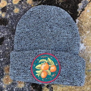 May include: Gray knitted beanie with a circular embroidered patch featuring three orange fruits, green leaves, and brown seeds on a green background. The patch has a red border.