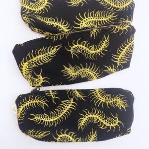 May include: Three black zippered pouches with a repeating yellow centipede pattern. The pouches are rectangular with rounded ends. The zippers are silver. The background is white.