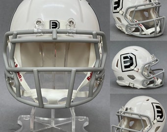 Op de afbeelding: Witte miniatuur voetbalhelm met een grijs gezichtsmasker en een zwart "D" logo aan de zijkant. De helm staat op een transparante kunststof standaard.