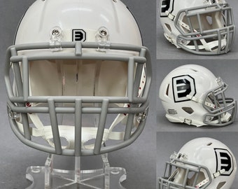 May include: White football helmet with a gray face mask and a black "D" logo. The helmet is on a clear acrylic display stand.