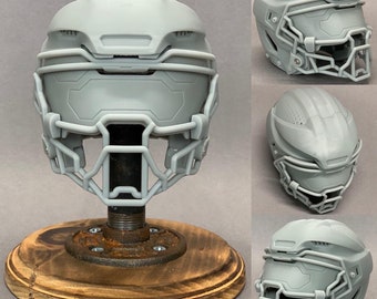 Op de afbeelding: Een grijze plastic voetbalhelm met een gezichtsmasker op een houten standaard. De helm heeft een getextureerd oppervlak en een vizier.