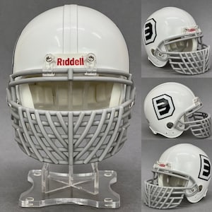 May include: White football helmet with a gray face mask and a clear plastic stand. The helmet has a black "E" on the side and the word "Riddell" printed on the front.