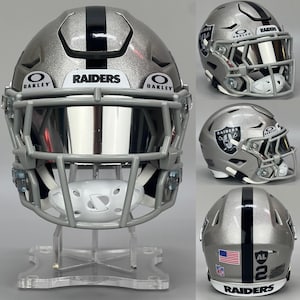 May include: Silver and black Raiders football helmet with a gray facemask. The helmet features the Raiders logo, the Oakley logo, and the text "RAIDERS". The back of the helmet displays the American flag and the number 2.