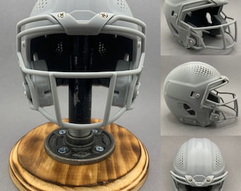 Op de afbeelding: Een grijze miniatuur American footballhelm met een gezichtsmasker, gemonteerd op een houten voet. De helm heeft een gedetailleerd ontwerp met ventilatiegaten en is bevestigd aan een zwarte buis. De voet is rond en gemaakt van lichtbruin hout.