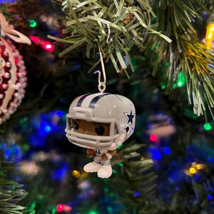 May include: A white and blue Christmas ornament featuring a football player wearing a helmet with a star on it. The ornament is hanging from a Christmas tree.