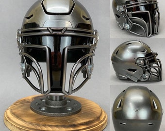 Op de afbeelding: Een zwarte voetbalhelm met een gezichtsmasker en een zwart vizier. De helm staat op een houten standaard.