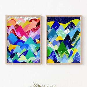 May include: Two framed art prints depicting abstract mountain scenes. The artwork uses a vibrant palette of blues, greens, pinks, and yellows, with white accents and a dotted pattern. The frames are a light wood colour.