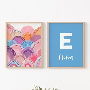May include: Two framed art prints. One features a watercolour design of layered, semi-circular shapes in shades of pink, orange, purple, and blue. The other has a light blue background with a white letter "E" and the name "Emma" in script.