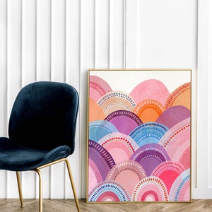 May include: A framed watercolour painting with an abstract landscape design. The artwork displays layered semi-circles in pink, purple, orange, and blue, with dotted and arched details. The frame is gold, and the painting is next to a blue velvet chair.