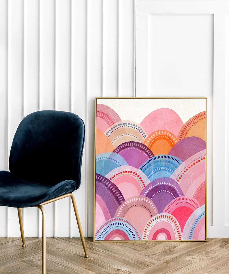 May include: A framed watercolour painting with an abstract landscape design. The artwork displays layered semi-circles in pink, purple, orange, and blue, with dotted and arched details. The frame is gold, and the painting is next to a blue velvet chair.
