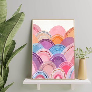 May include: A framed watercolour print showcasing an abstract landscape. The artwork presents layered, semi-circular forms in pink, purple, orange, and blue, with dotted and arched details. The print is displayed on a white shelf, with a plant in the foreground.