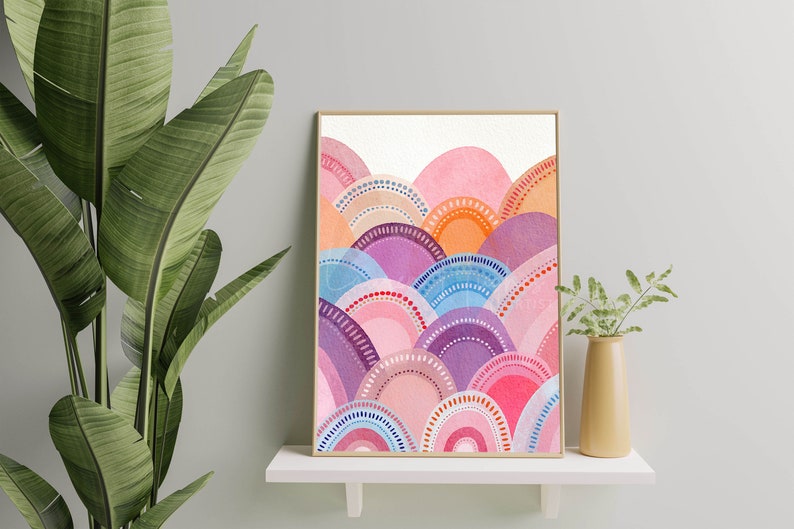 May include: A framed watercolour print showcasing an abstract landscape. The artwork presents layered, semi-circular forms in pink, purple, orange, and blue, with dotted and arched details. The print is displayed on a white shelf, with a plant in the foreground.