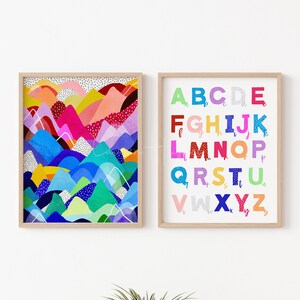 May include: Two framed art prints. One depicts a colourful abstract landscape with mountains in shades of blue, pink, yellow, and green. The other print shows the alphabet in various colours and patterns. Both are in light wood frames.